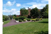 Green lawn and landscaped pathway leading to a gazebo and a multi-story brick building under a partly cloudy sky.