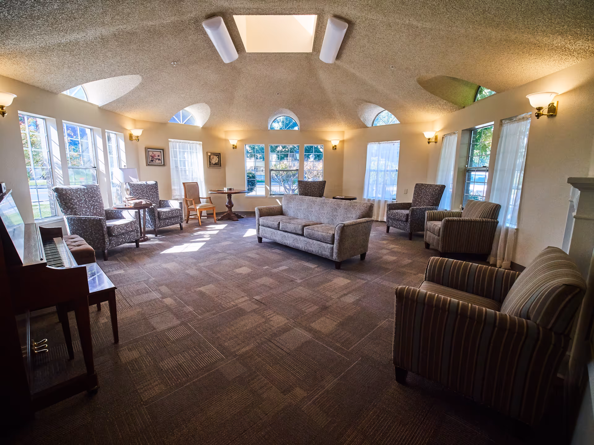 A spacious, well-lit living room with multiple armchairs and a sofa arranged around the room. Large windows with sheer curtains allow natural light to fill the space. The ceiling has a skylight and several wall-mounted light fixtures. A piano is visible on the left side of the room.