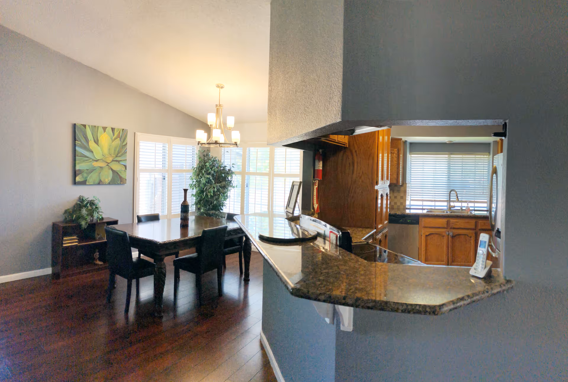 Interior view of a senior care home showing a dining area with a wooden table and six black chairs, a chandelier above the table, a large window with white blinds, a green plant, and a painting on the wall. Adjacent to the dining area is a kitchen with wooden cabinets, a granite countertop, a sink under a window with blinds, and a cordless phone on the counter.