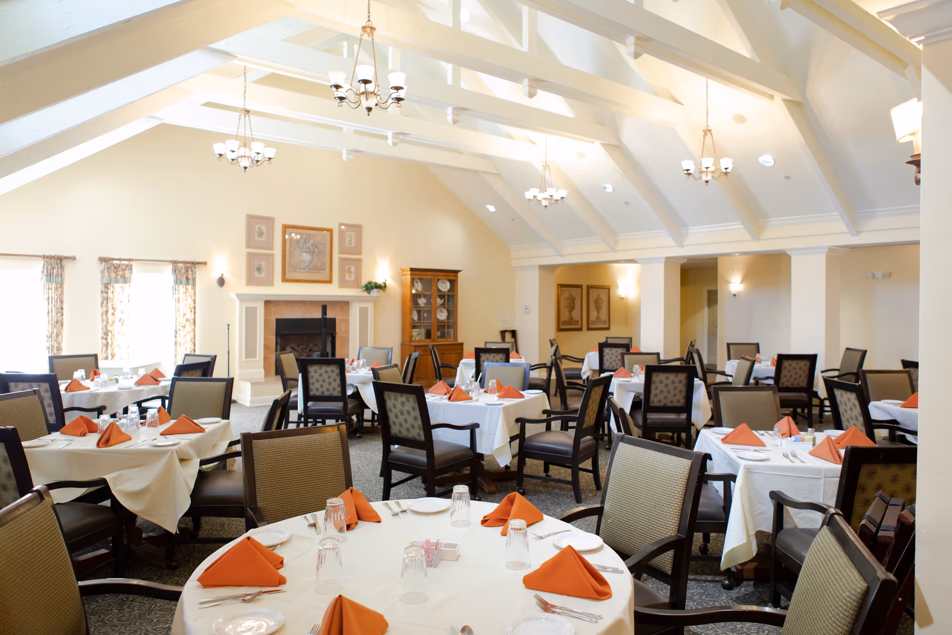 A spacious dining room with multiple round and square tables covered with white tablecloths and set with orange folded napkins, plates, glasses, and silverware. The room features high vaulted ceilings with exposed white beams and several chandeliers. There is a fireplace with framed artwork above it and a wooden cabinet displaying dishes against the far wall. The room is well-lit with natural light coming through windows with floral curtains.