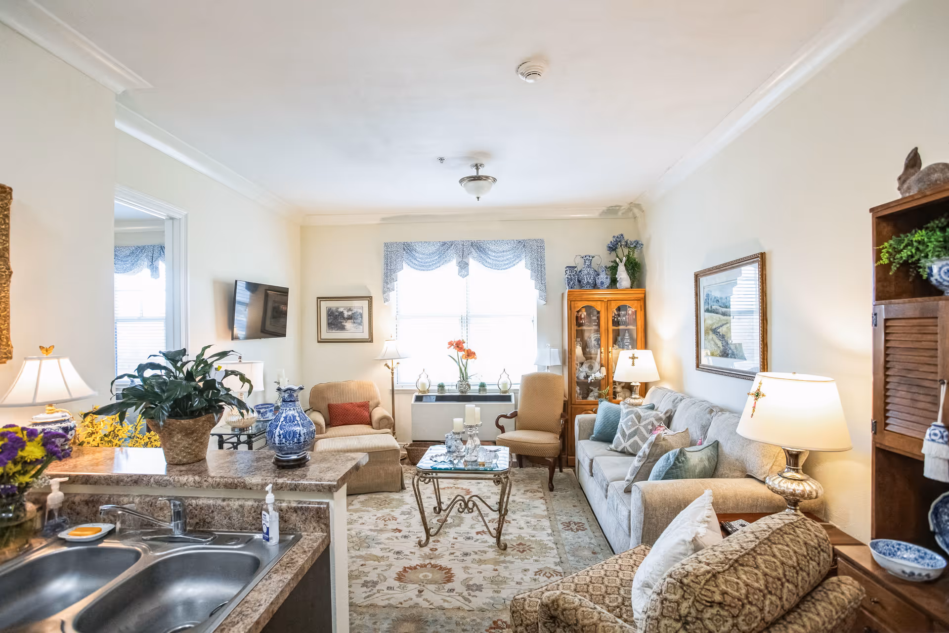 A cozy living room with a beige sofa, armchairs, a glass coffee table, and a patterned rug. The room is decorated with lamps, plants, blue and white vases, and framed artwork. A kitchen sink and counter with a potted plant and hand sanitizer are visible in the foreground.
