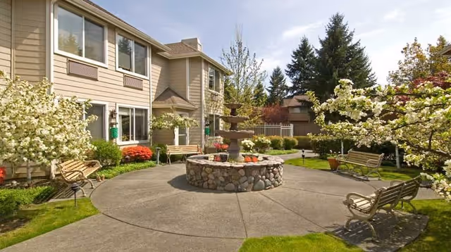 Outdoor courtyard area of a senior living facility with a stone fountain in the center, surrounded by benches and flowering trees. The building exterior is beige with multiple windows, and there is a well-maintained lawn and garden area.
