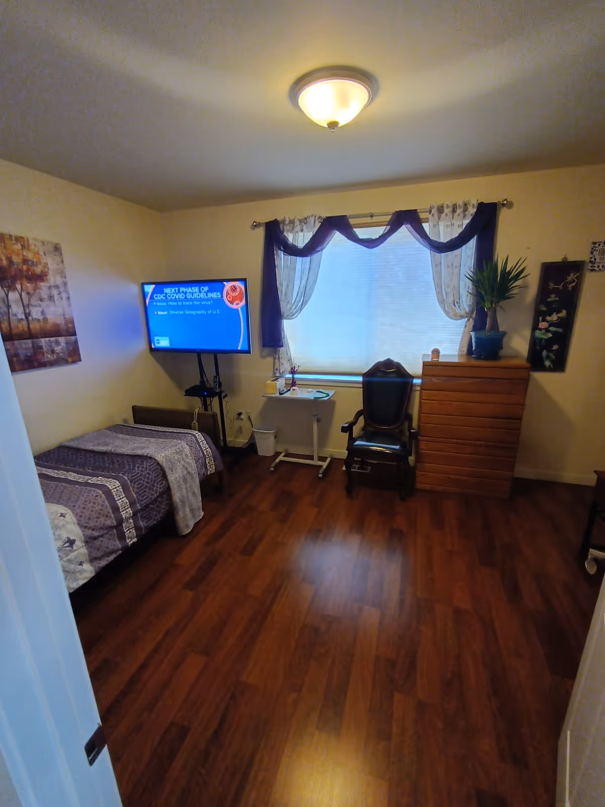 A small bedroom with a single bed covered in a purple and white patterned blanket, a flat-screen TV mounted on a stand displaying CDC COVID guidelines, a window with sheer and purple curtains, a wooden dresser with a potted plant on top, a black chair, and a small white table with a cup and some items on it. The room has wooden flooring and a ceiling light fixture.