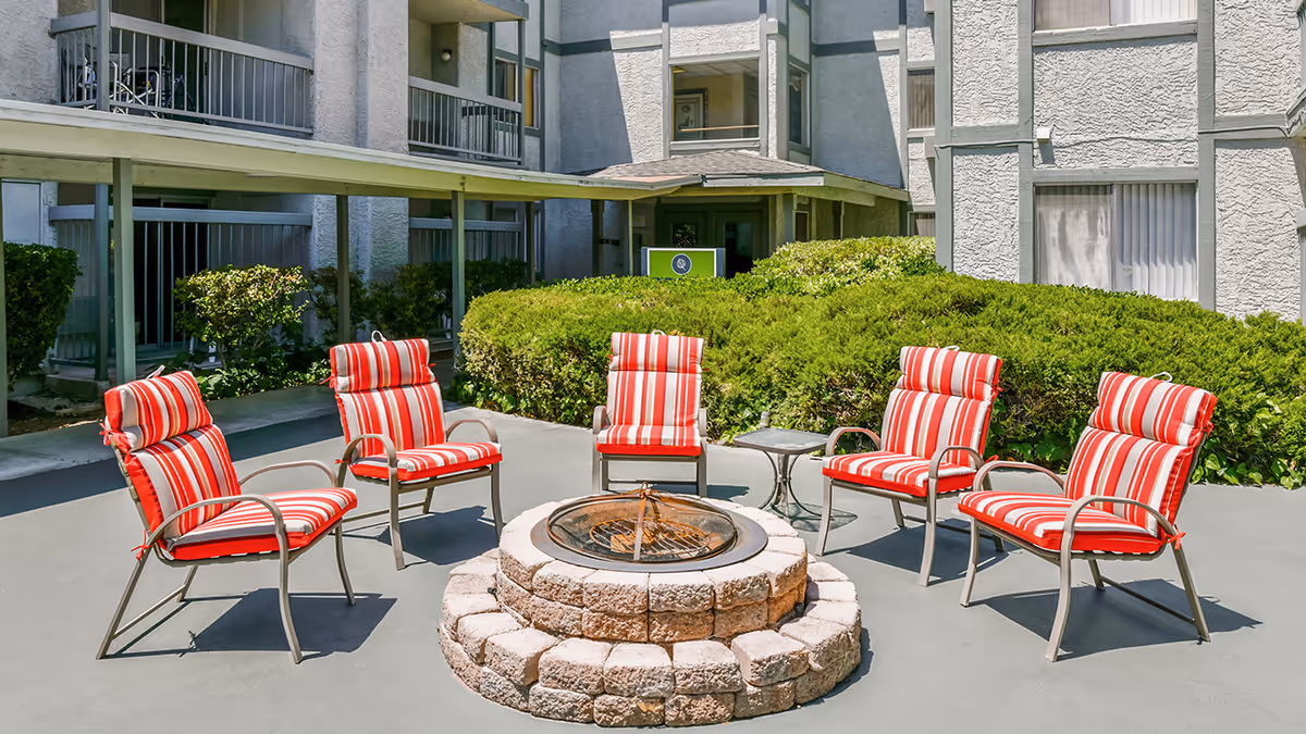 Outdoor patio area at Holiday Montara Meadows with five metal chairs featuring red and white striped cushions arranged around a circular stone fire pit. The background shows the exterior of the building with windows, balconies, and green shrubbery.