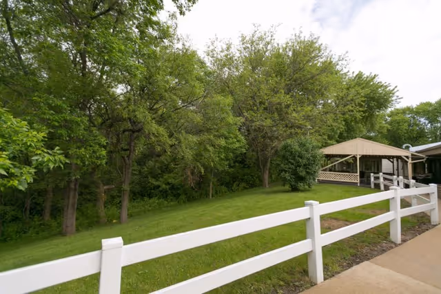 White split-rail fence along a grassy lawn with trees and a covered gazebo next to a building under a cloudy sky.