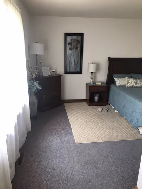 Bright bedroom with a bed, nightstand and dresser beside a curtained window and framed wall art.