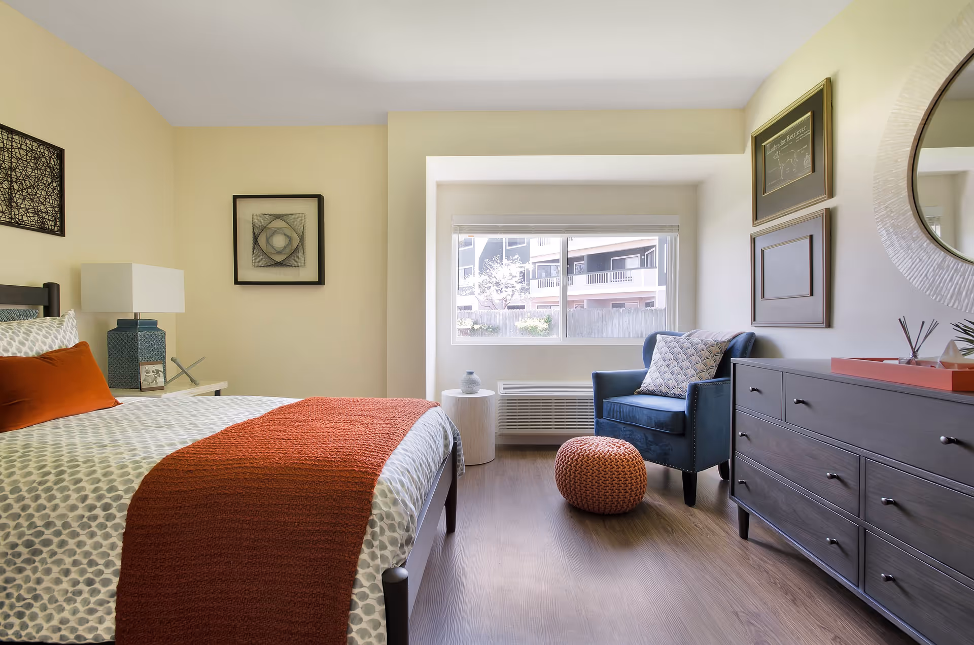 A bright and cozy bedroom with a bed featuring white and gray patterned bedding and an orange throw blanket. Next to the bed is a nightstand with a blue lamp and decorative items. Across from the bed is a dark wooden dresser with a round mirror above it and framed artwork on the wall. A blue armchair with a patterned pillow and an orange knitted pouf are placed near a window letting in natural light.