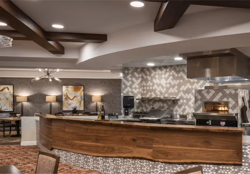 Interior view of a senior living facility kitchen area with a wooden counter in the foreground, tiled backsplash, and stainless steel cooking appliances including a pizza oven. In the background, there is a dining area with tables, chairs, lamps, and abstract wall art.