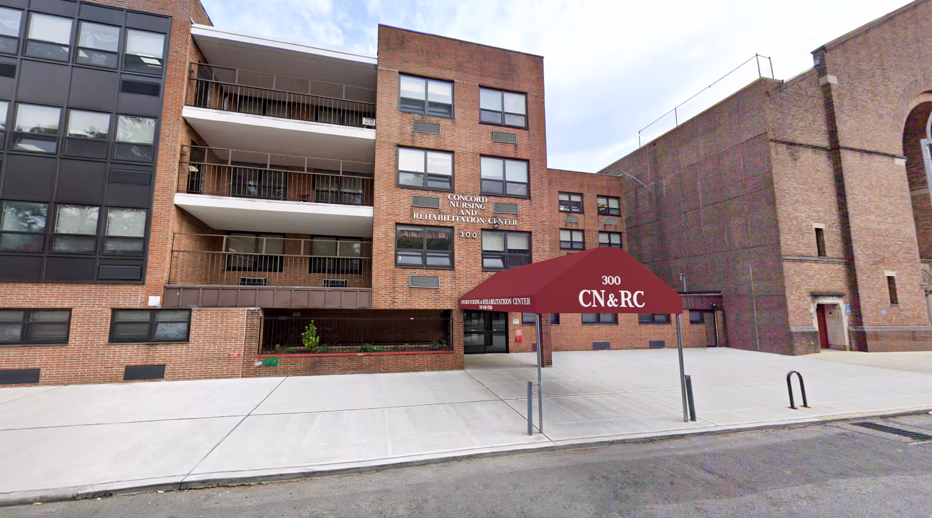 Exterior view of Concord Nursing and Rehabilitation Center, a multi-story brick building with balconies and large windows. There is a maroon canopy over the entrance with the text '300 CN&RC'. The sidewalk and street are visible in front of the building.