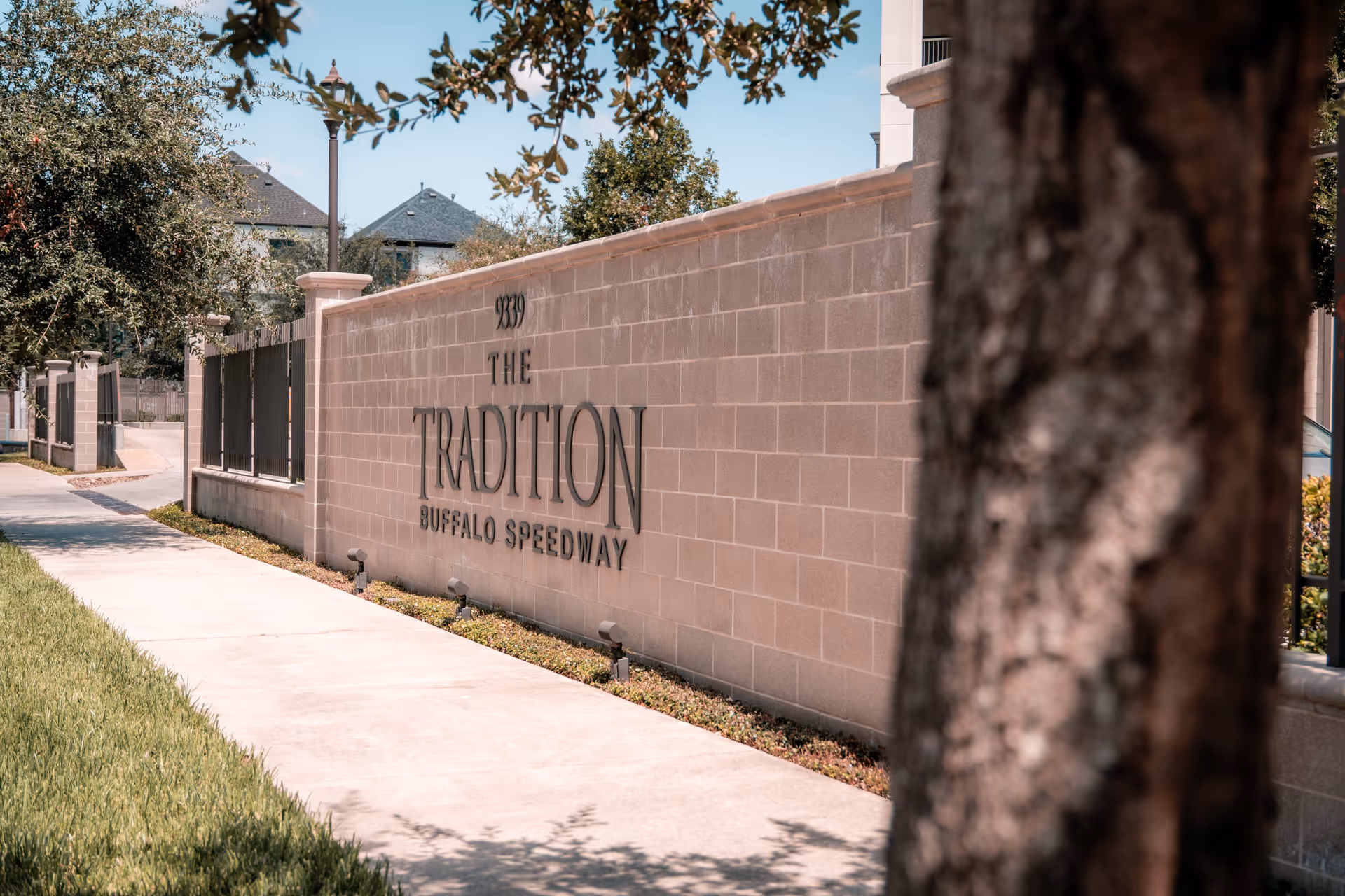 A sidewalk running alongside a beige brick wall with the text '9339 THE TRADITION BUFFALO SPEEDWAY' mounted on it. There are trees and a lamp post along the sidewalk, with residential buildings visible in the background under a clear blue sky.