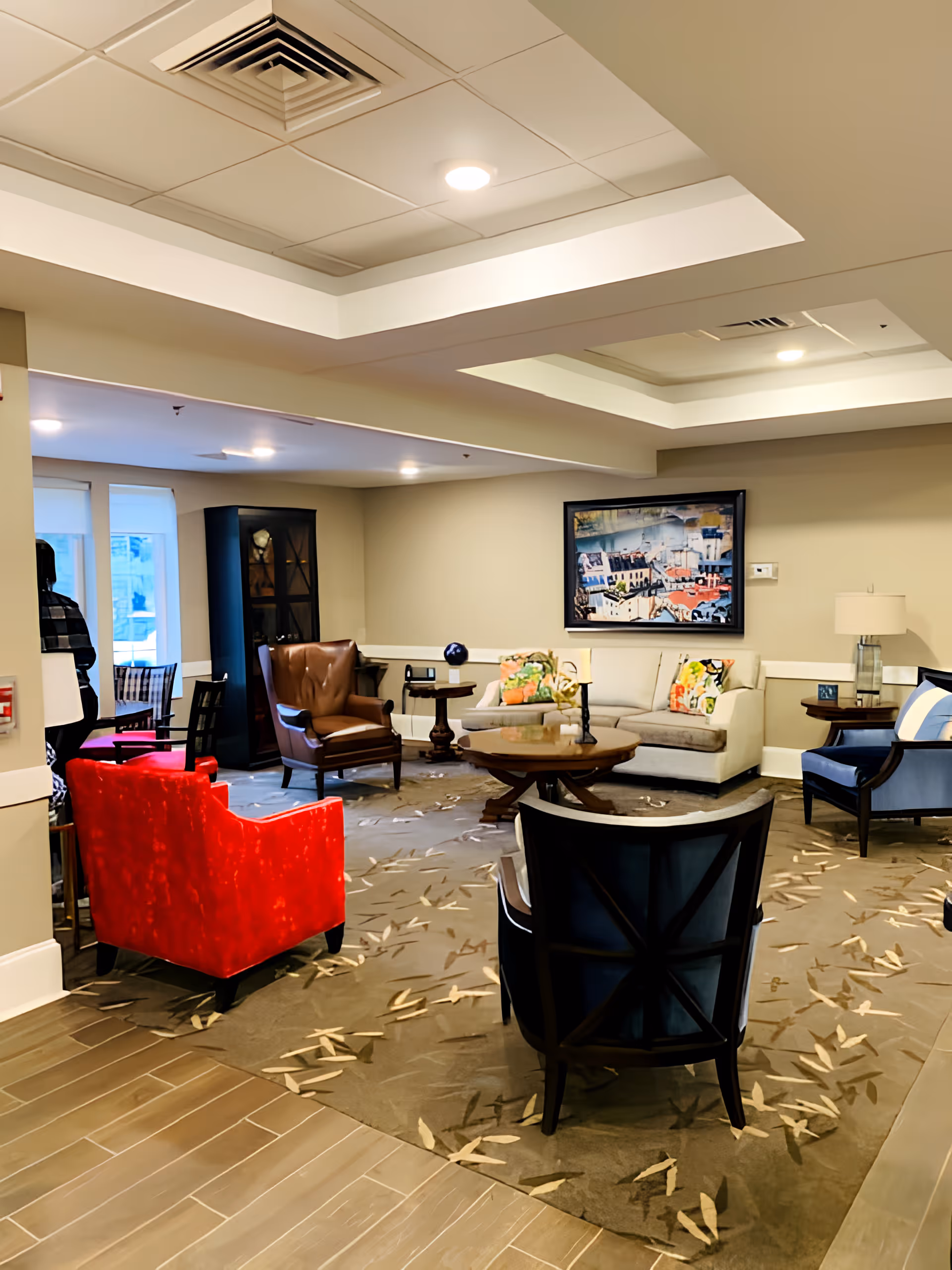A cozy living room area in a senior living facility with a variety of chairs including a bright red armchair, a brown leather armchair, and blue cushioned chairs arranged around a wooden coffee table on a patterned carpet. A beige sofa with colorful pillows is against the wall beneath a framed artwork. The ceiling has recessed lighting and a decorative tray design.