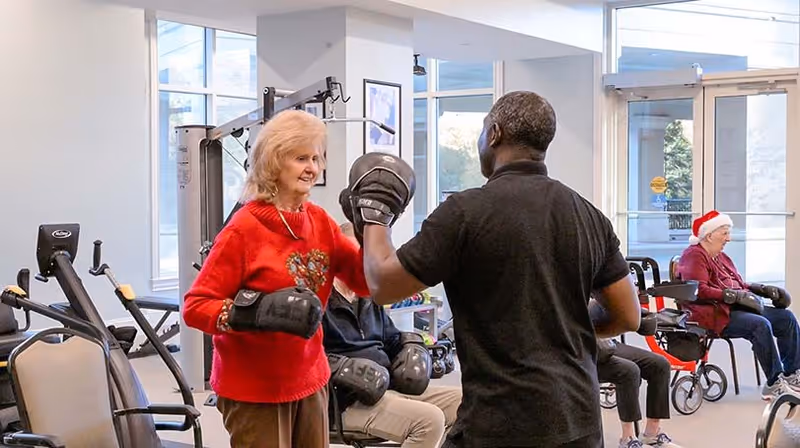 An elderly woman wearing a red sweater with a heart design is practicing boxing with a trainer holding boxing pads in a fitness room. Other seniors are seated in the background, some wearing boxing gloves, and one person is wearing a Santa hat. Exercise equipment and large windows are visible in the room.