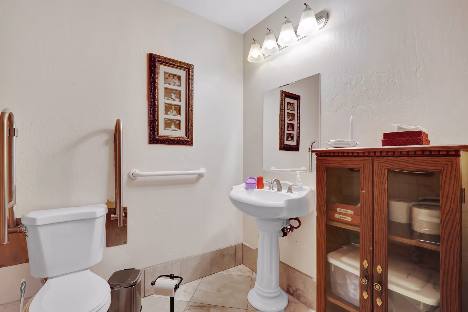Accessible bathroom with a toilet and grab bars, a pedestal sink under a mirror, and a wooden storage cabinet.