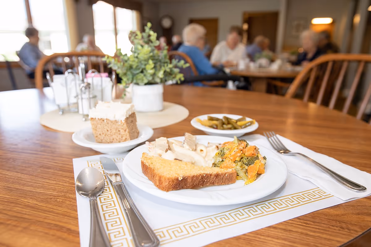 A close-up view of a meal on a wooden dining table in a senior living facility. The plate contains a slice of cornbread, pasta with white sauce, and mixed vegetables. There is also a small plate with green beans and a piece of cake with white frosting. In the background, several elderly people are seated and engaged in conversation in a well-lit dining room.