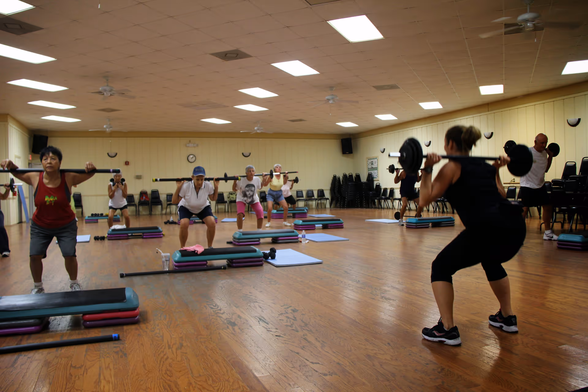 A group of people participating in a fitness class in a large room with wooden floors and beige walls. They are performing squats while holding weighted bars across their shoulders. Exercise steps and mats are placed on the floor in front of them.