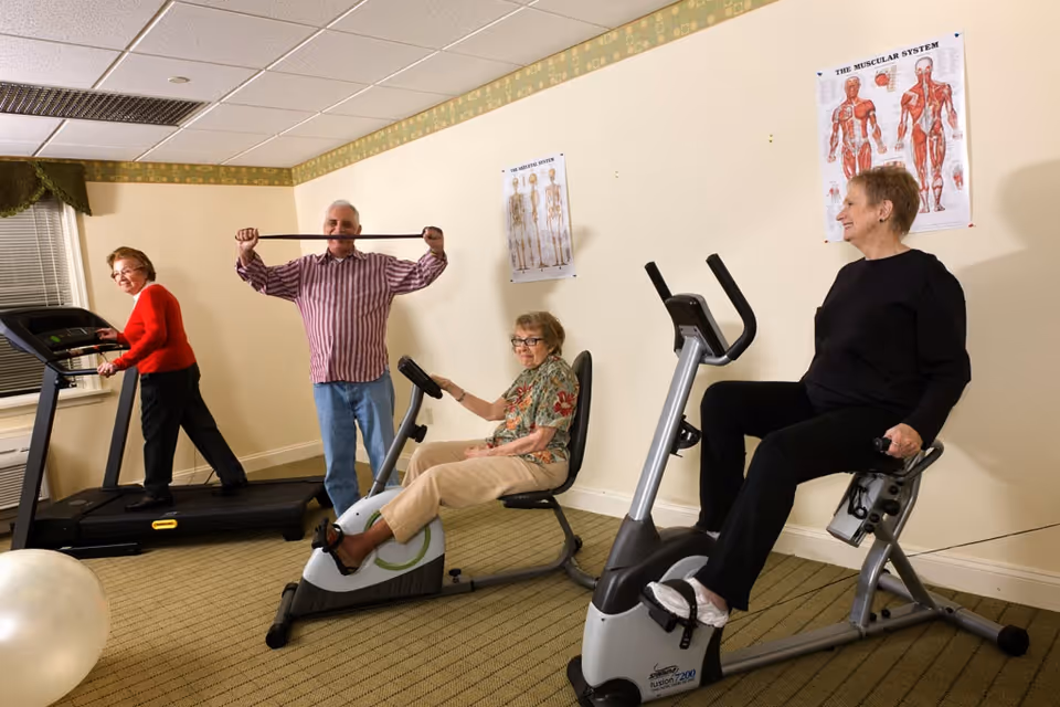 Four elderly people exercising in a fitness room. One woman is walking on a treadmill, a man is stretching with a resistance band, and two women are seated on stationary exercise bikes. The room has beige walls with anatomical posters and a carpeted floor.