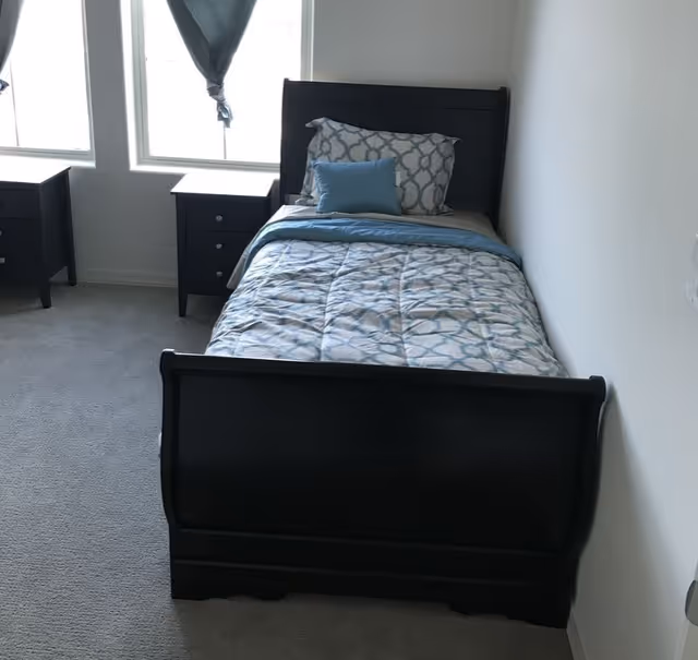 Small bedroom with a twin bed dressed in blue patterned bedding, two dark nightstands and windows on the left.