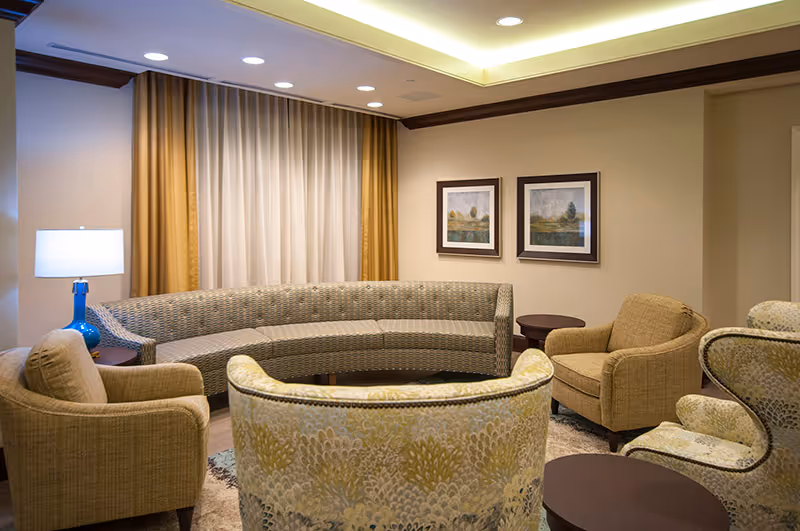 A cozy living room area with a curved sofa, several upholstered armchairs, a blue table lamp on a side table, two framed landscape paintings on the wall, and beige curtains covering a window.