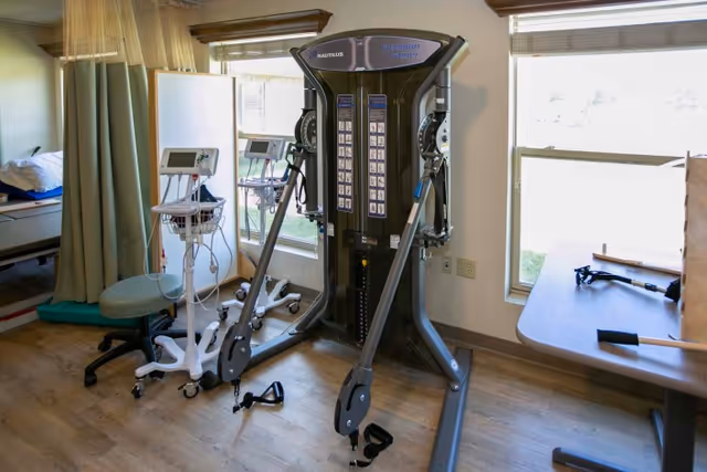 A rehabilitation or physical therapy room with exercise equipment including a Nautilus Freedom Trainer machine, medical monitors on wheeled stands, a green stool, a hospital bed partially visible behind a curtain, and a table near a window with blinds.