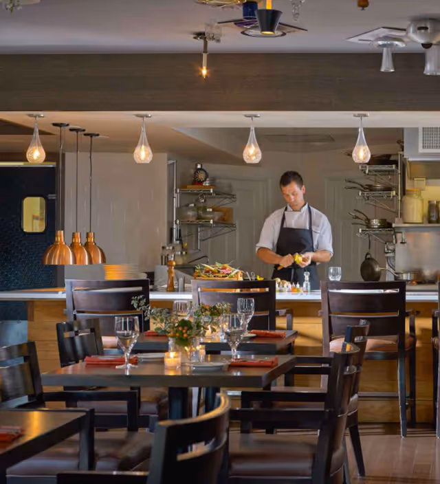 Dining room with set tables and a chef preparing food behind a counter under hanging pendant lights.
