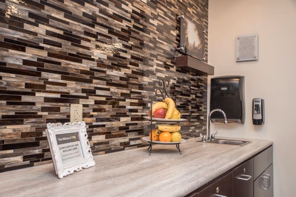 Small kitchenette counter with a mosaic tile backsplash, a two-tier fruit stand, sink, and wall-mounted soap and paper towel dispensers.