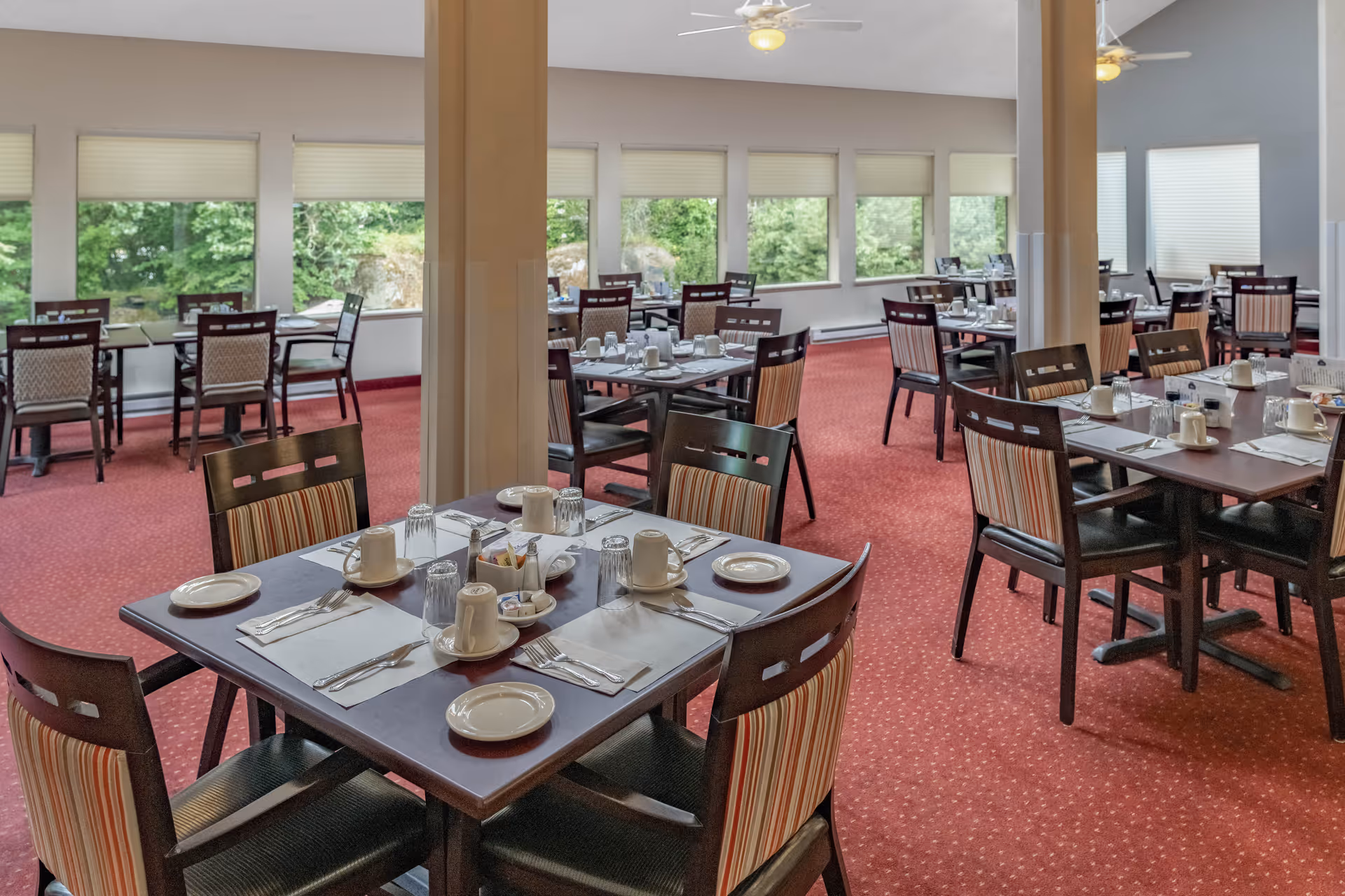 Dining room with multiple tables set for meals, upholstered chairs, red carpet, and large windows showing greenery outside.