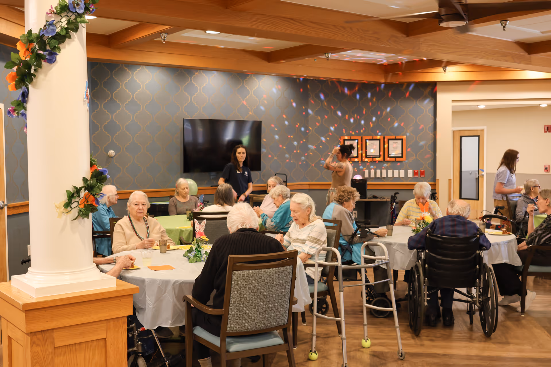 Elderly residents and staff sit around decorated tables in a communal dining/activity room with a TV and a flower-wrapped column.
