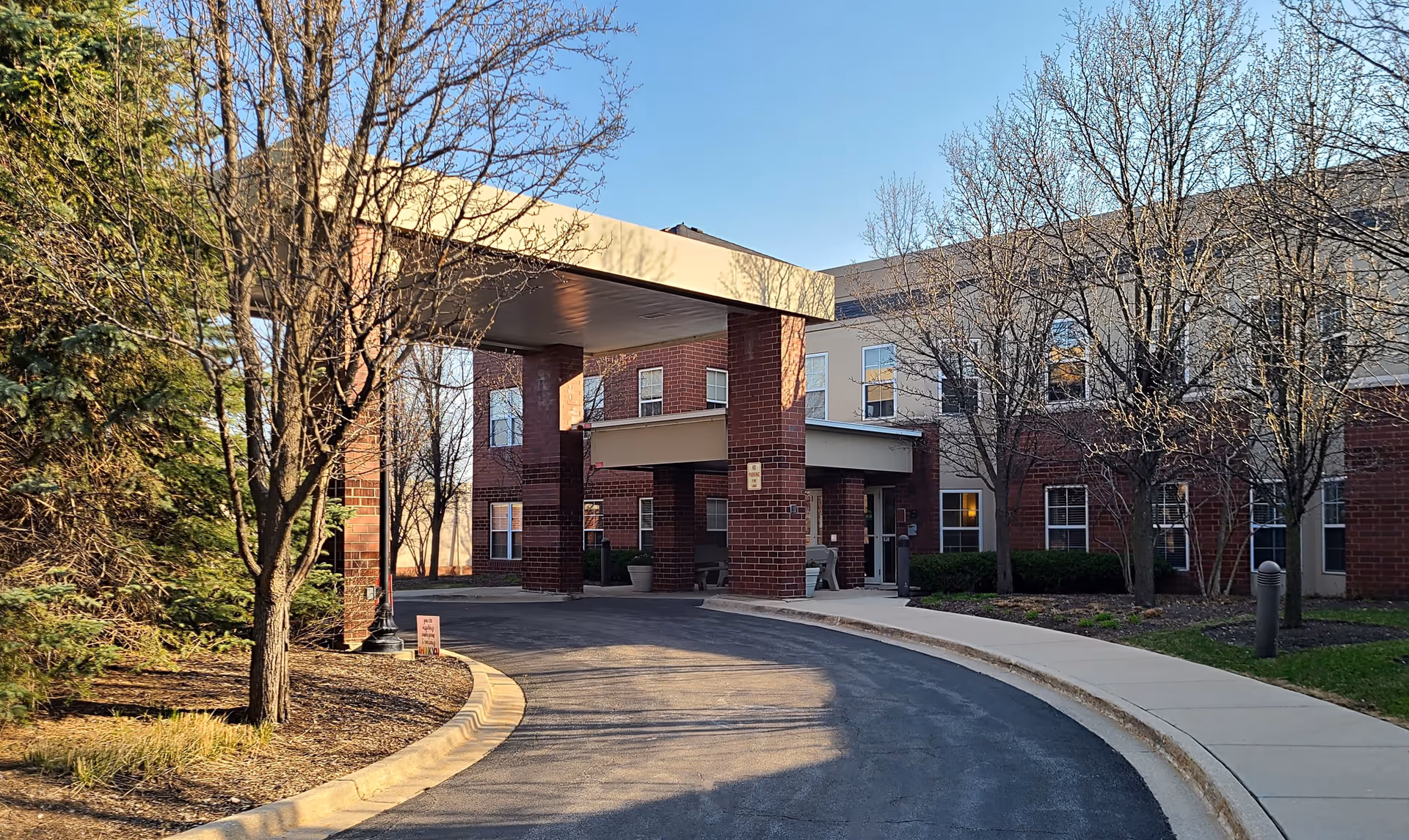 Entrance of a brick and beige building with a covered drop-off area, surrounded by leafless trees and a curved driveway under a clear blue sky.
