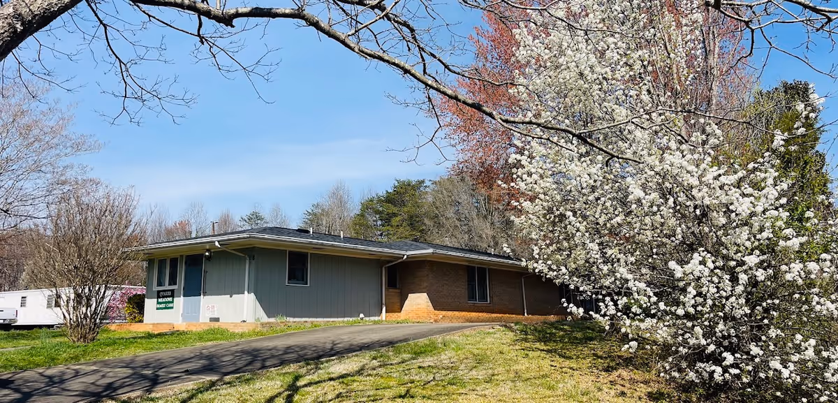 Single-story assisted living building with a driveway, trees, and blooming white blossoms nearby.