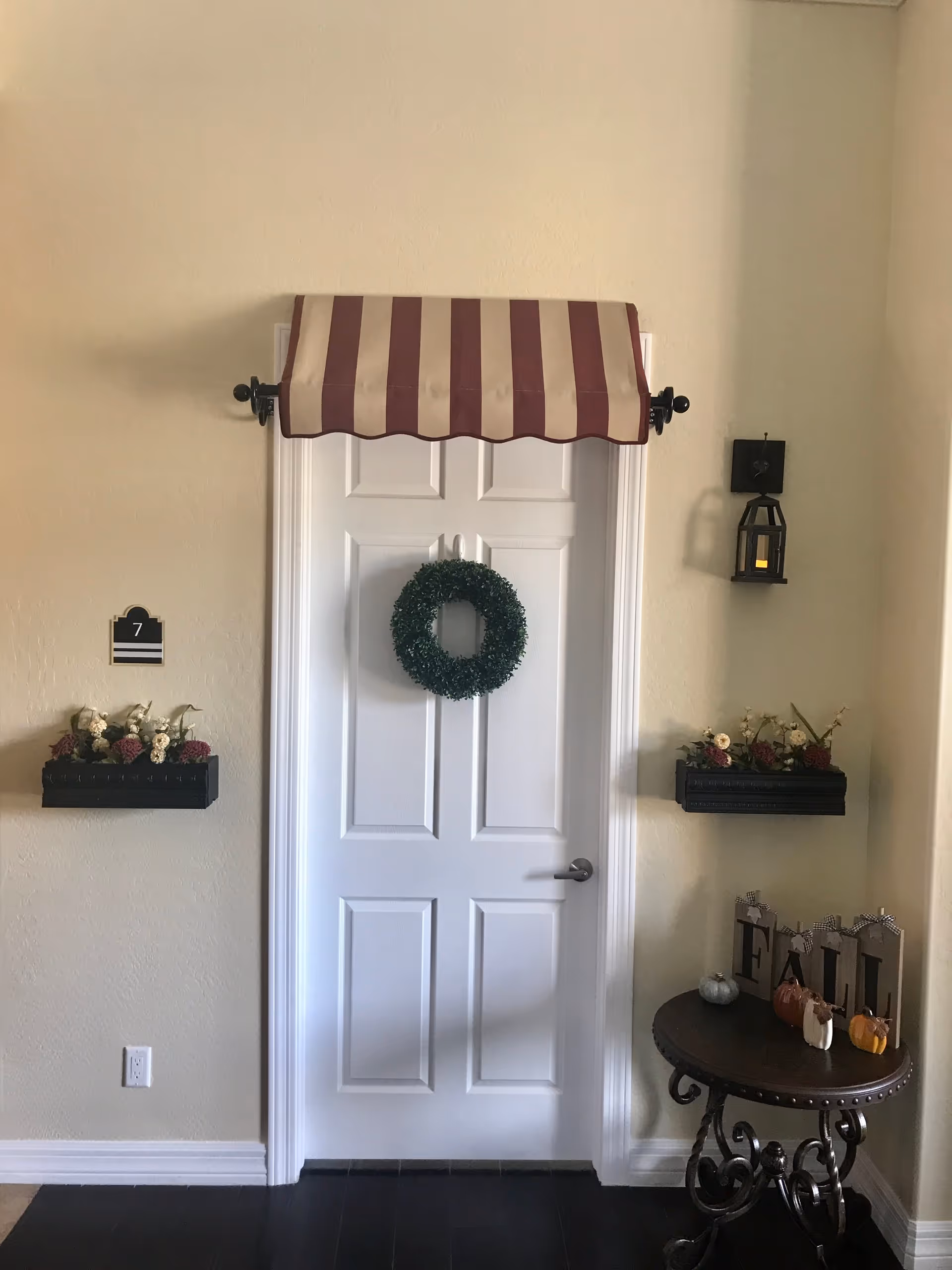 A white door with a green wreath hanging on it, topped by a red and beige striped awning. On the left side of the door is a black wall-mounted flower box with white and purple flowers and a plaque with the number 7. On the right side is a matching flower box and a black lantern-style wall light. Below the flower box on the right is a round wooden table with decorative fall-themed items including small pumpkins and blocks spelling 'FALL'.