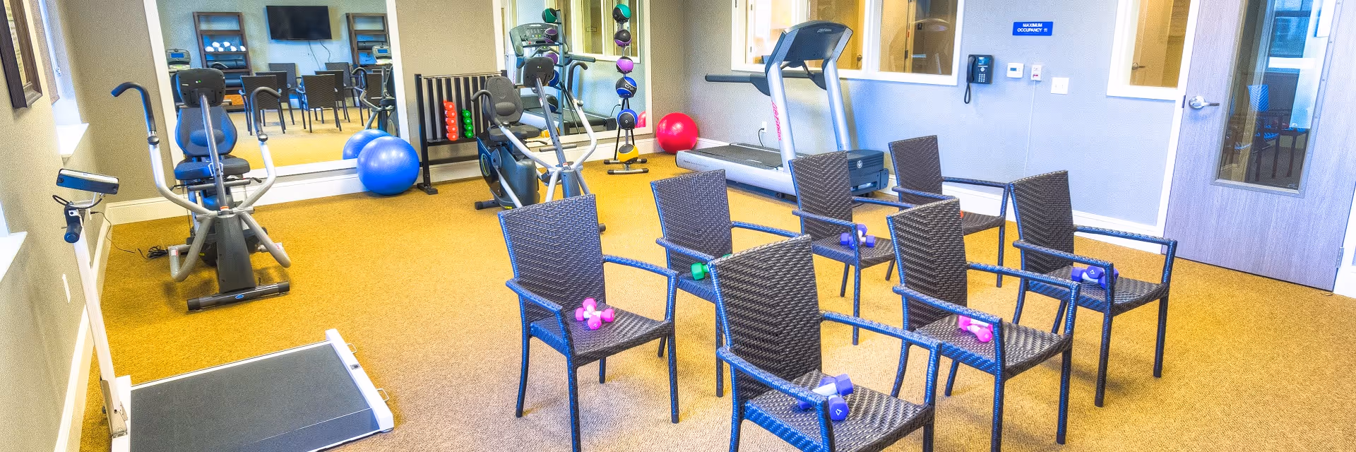 A senior living facility exercise room with six wicker chairs arranged in two rows, each chair holding a pair of small dumbbells. The room also contains exercise equipment including a treadmill, stationary bikes, an elliptical machine, exercise balls, and a rack of colorful dumbbells. A large mirror covers one wall, reflecting the equipment and chairs. There is a door with a window and a sign indicating maximum occupancy on the wall.