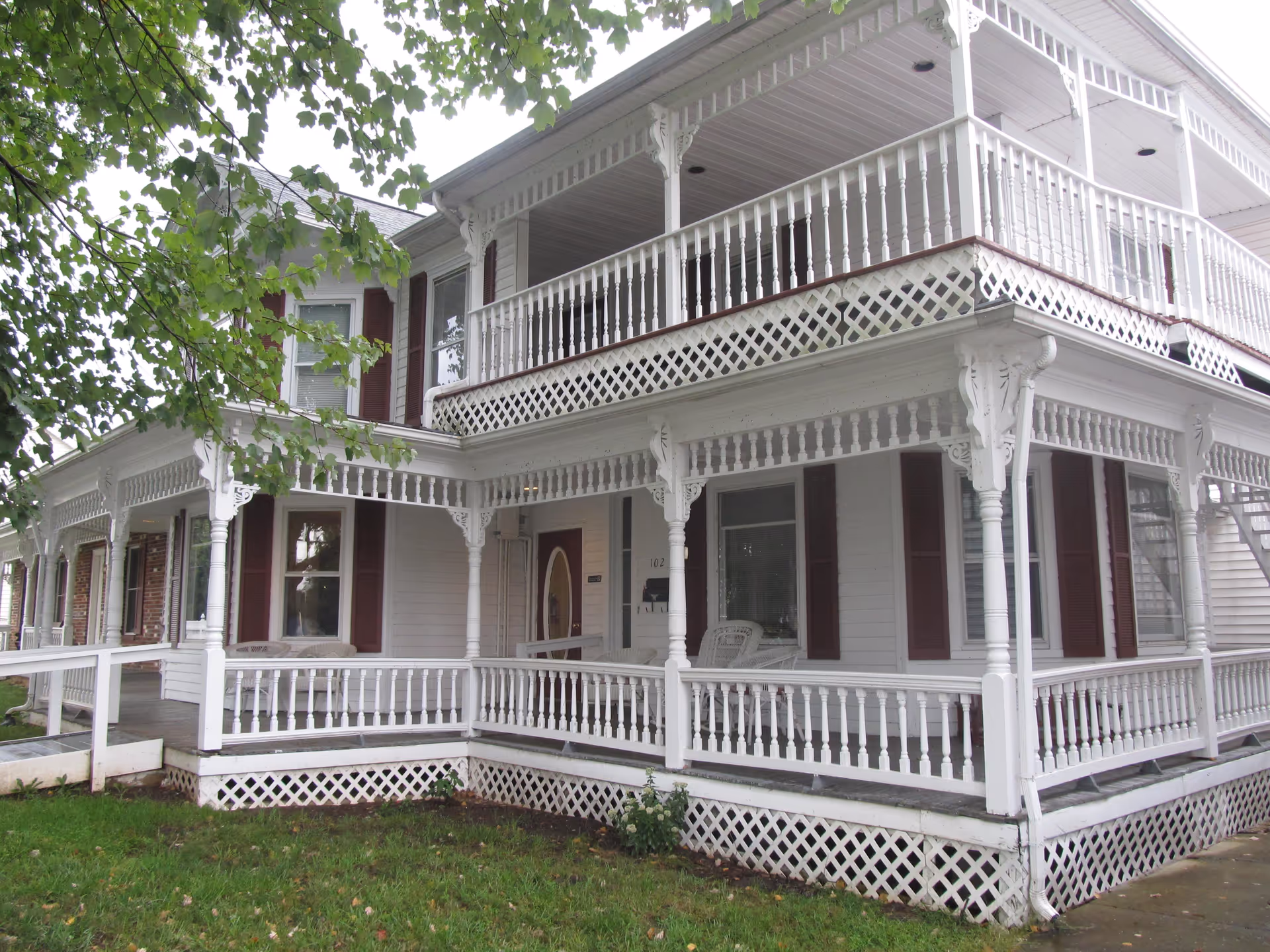 Exterior view of a two-story white house with a wraparound porch featuring decorative railings and columns. The house has maroon shutters on the windows and a green lawn in the foreground with a tree partially visible on the left side.