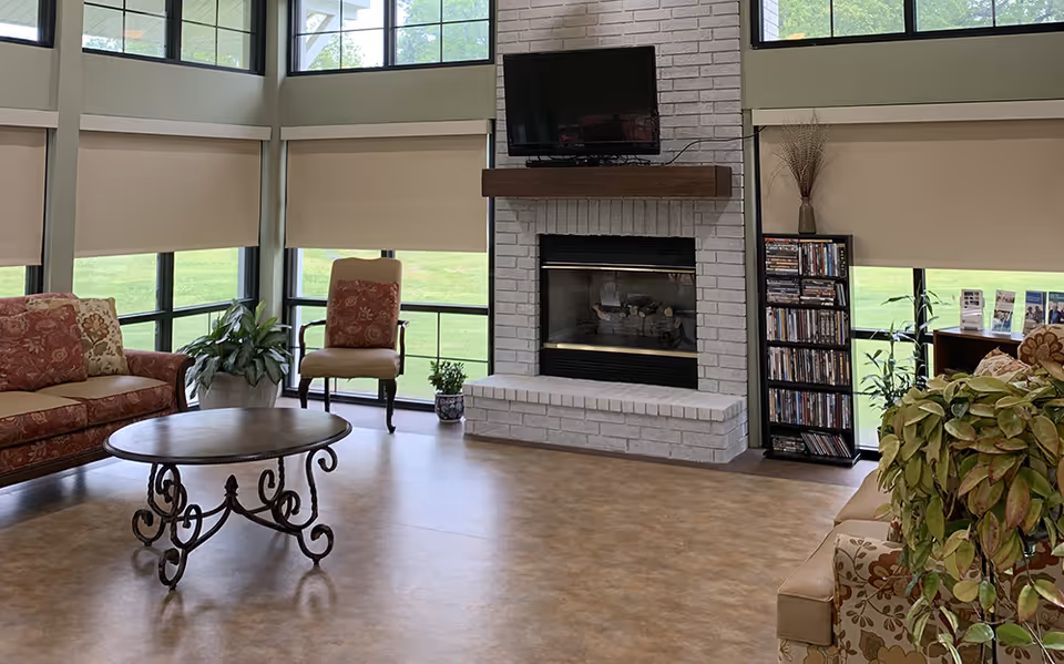 Bright common room with large windows, a central brick fireplace and TV, sofas, chairs, a coffee table, and plants.