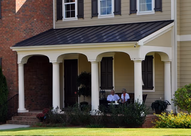 Two elderly people sitting and relaxing on a covered front porch of a house with white columns and beige siding, surrounded by greenery and a well-maintained lawn.