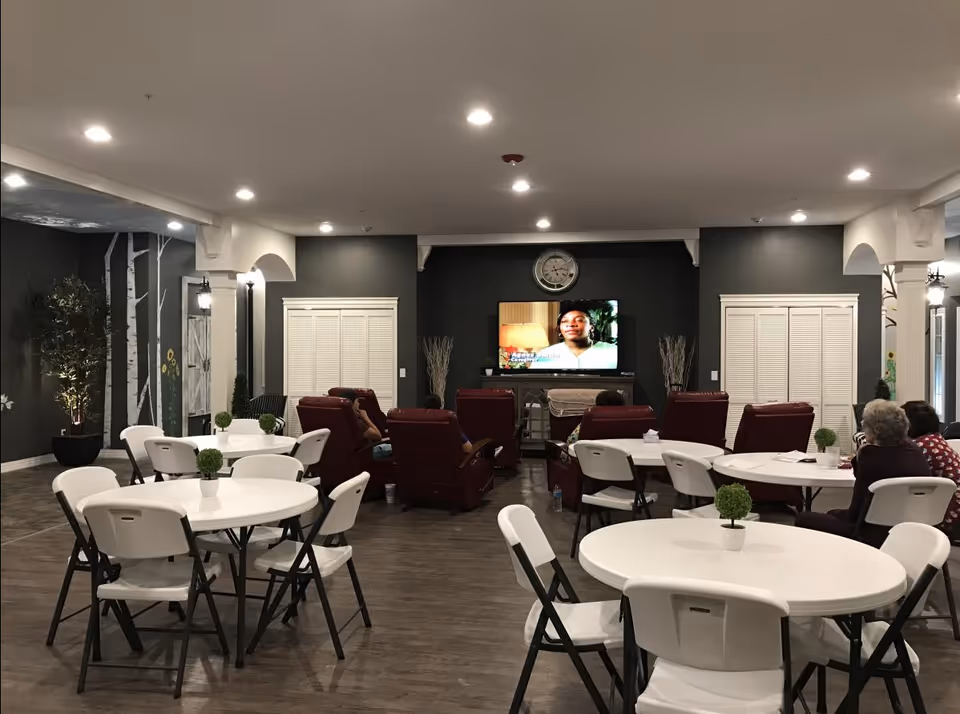 Senior living common room with round tables and white folding chairs, red recliners facing a wall-mounted TV beneath a wall clock.