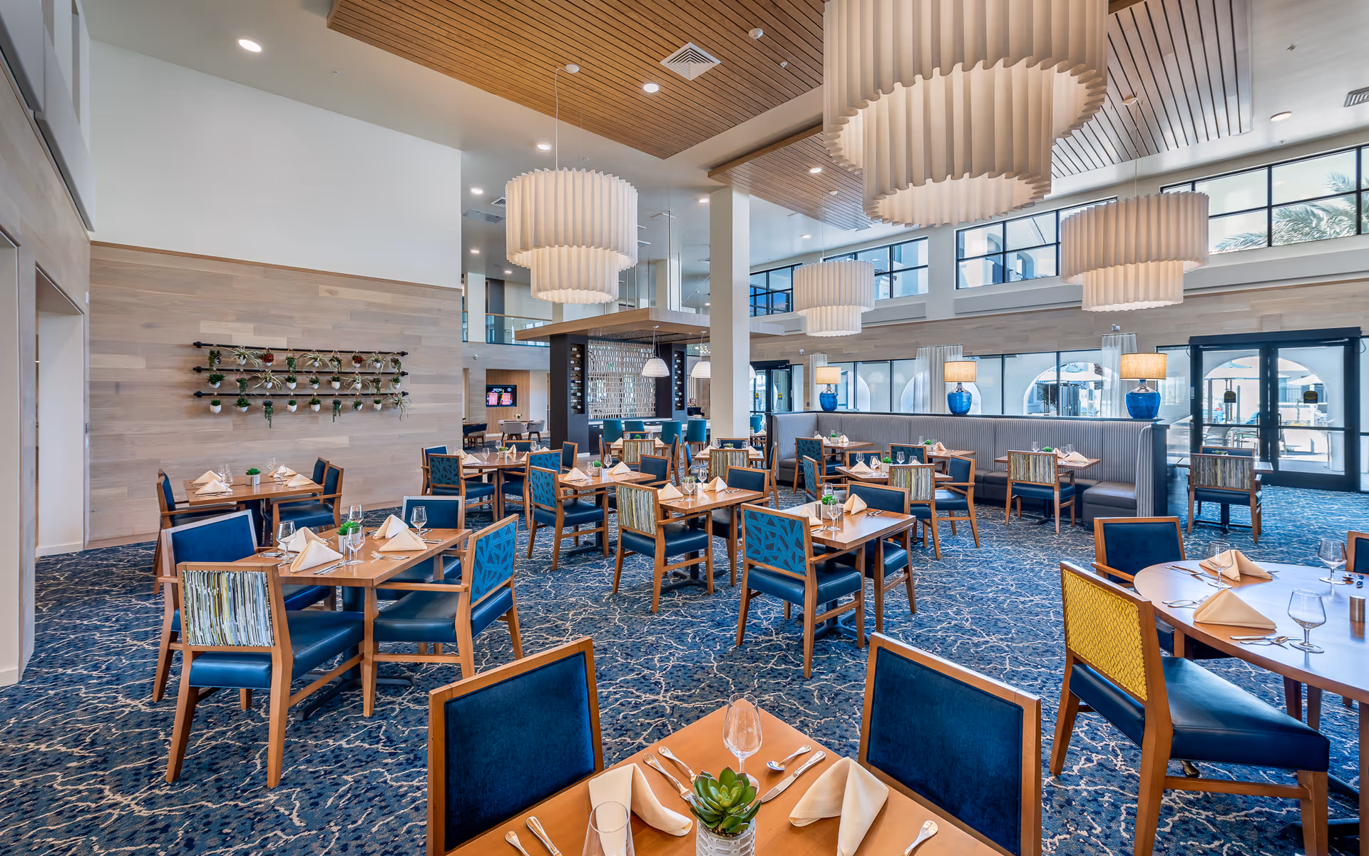 A spacious and modern dining room with multiple wooden tables set with folded napkins, glasses, and silverware. The chairs have blue upholstery with some featuring patterned backs. Large, elegant pendant lights hang from a wooden slatted ceiling. The room has large windows allowing natural light to fill the space, and there are blue vases with lamps on a long bench along one wall.