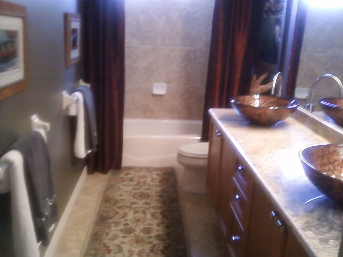 Bathroom with a bathtub, toilet, double vanity with vessel sinks, mirror, and a patterned rug.