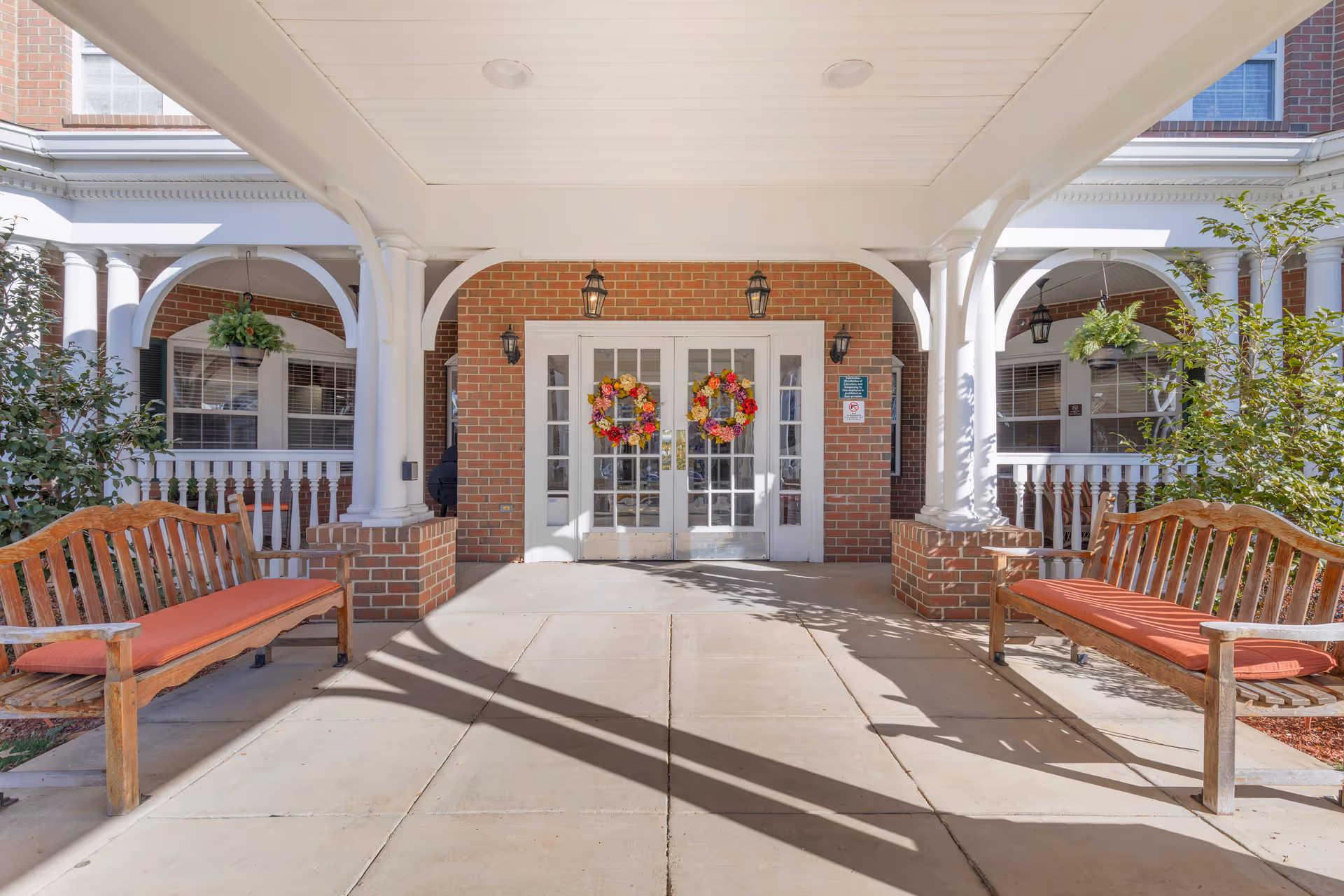 Covered entrance area of a brick building with white double doors decorated with colorful wreaths. Two wooden benches with orange cushions are placed on either side of the entrance. White columns and hanging plants are visible under the covered porch.