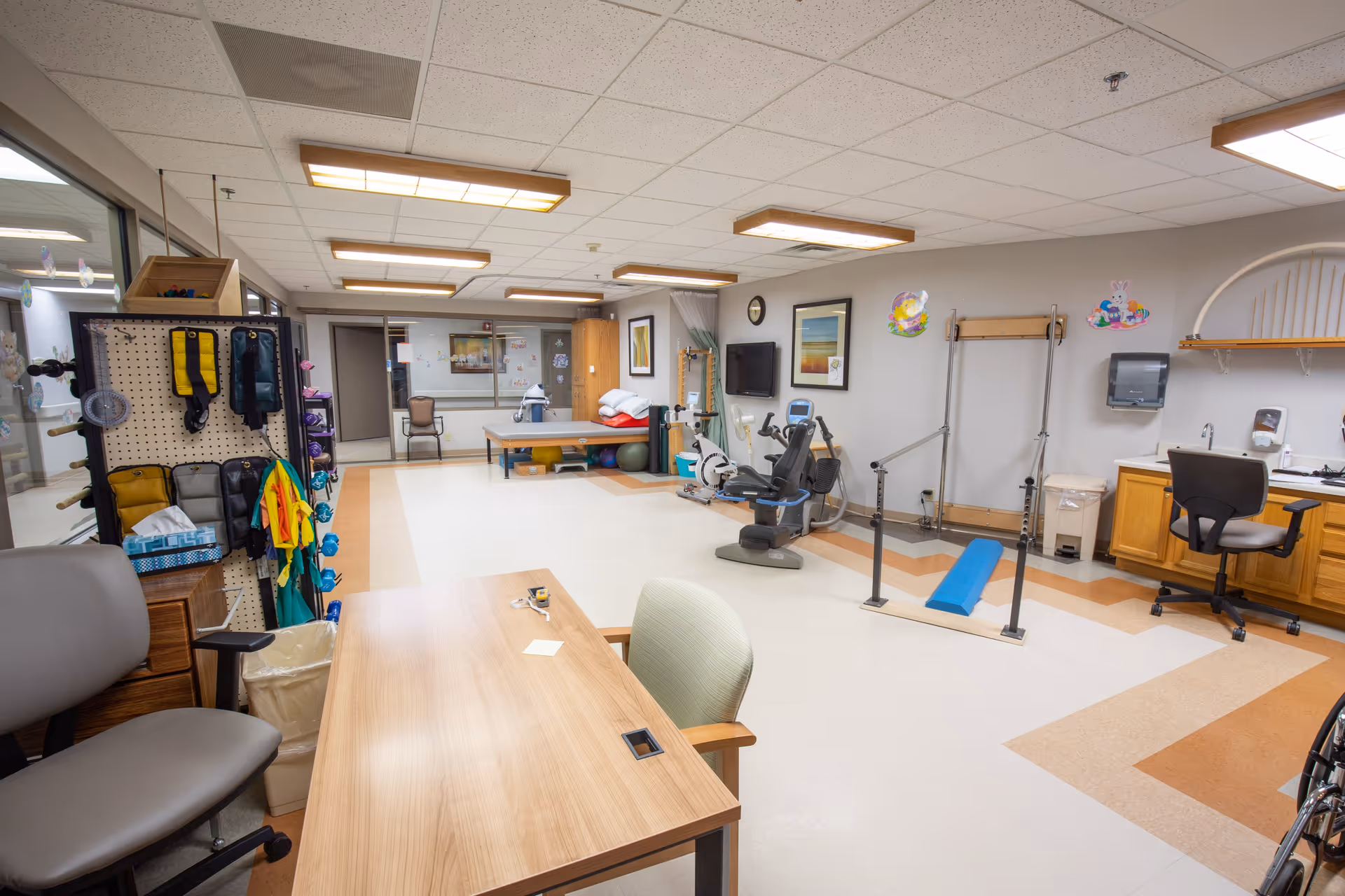 A spacious therapy or rehabilitation room in a healthcare facility with exercise equipment, parallel bars, a therapy table, chairs, and a desk. The room has bright overhead lighting, a wall-mounted TV, and various therapy tools and supplies organized around the space.