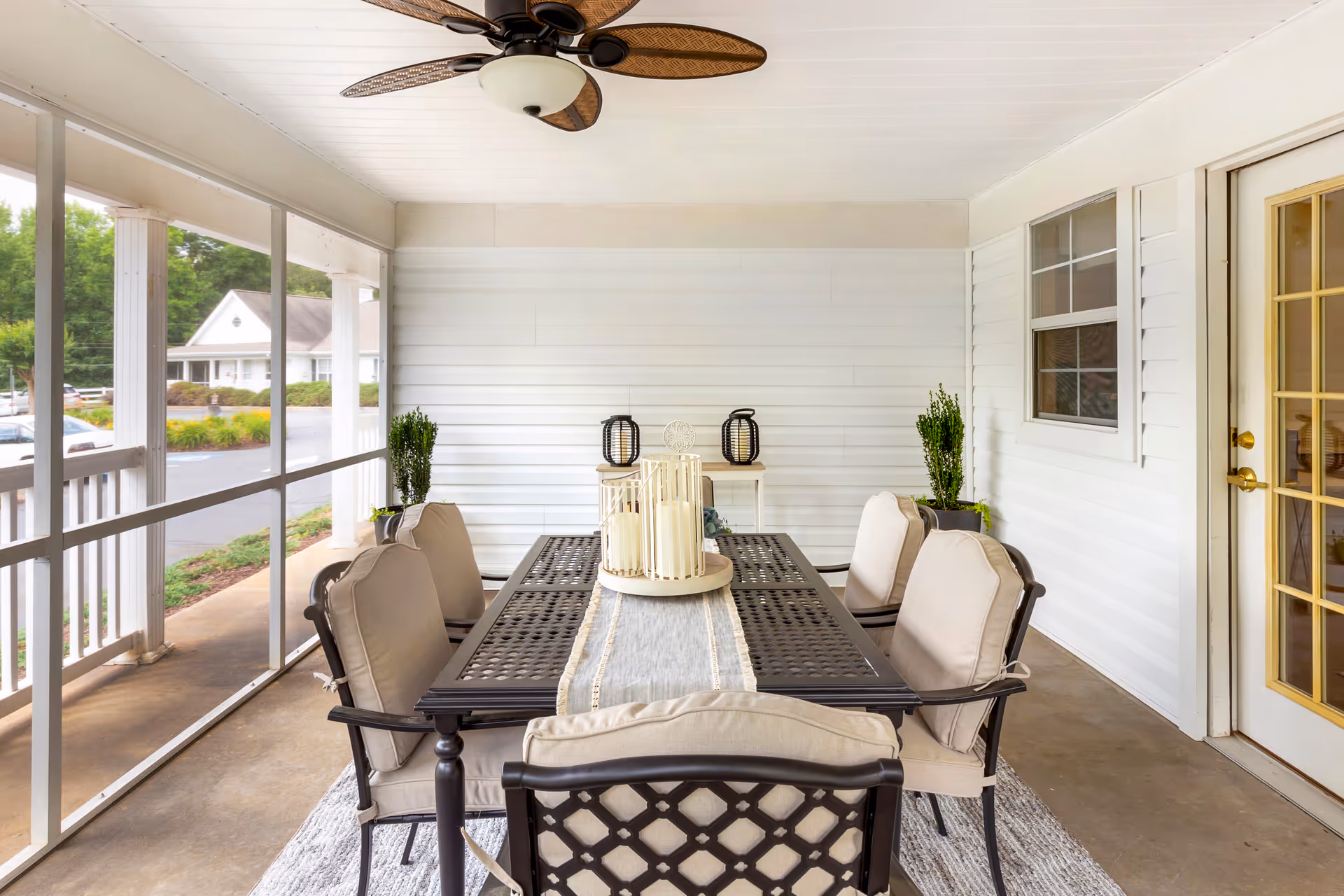A screened-in porch with a black metal dining table and six cushioned chairs. The table has a decorative centerpiece with candles. There are two potted plants on either side against a white paneled wall, a ceiling fan with leaf-shaped blades above, and a glass door leading inside. Outside the porch, a parking lot and greenery are visible.