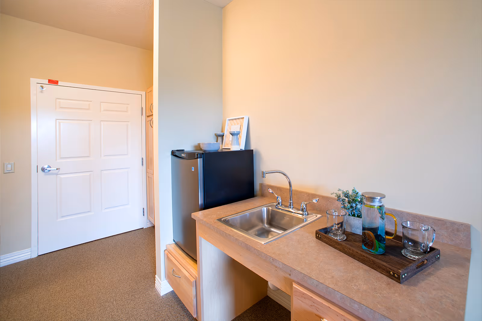 Small kitchenette with a countertop sink, mini fridge, and a tray holding a pitcher and glasses beside a closed white door.