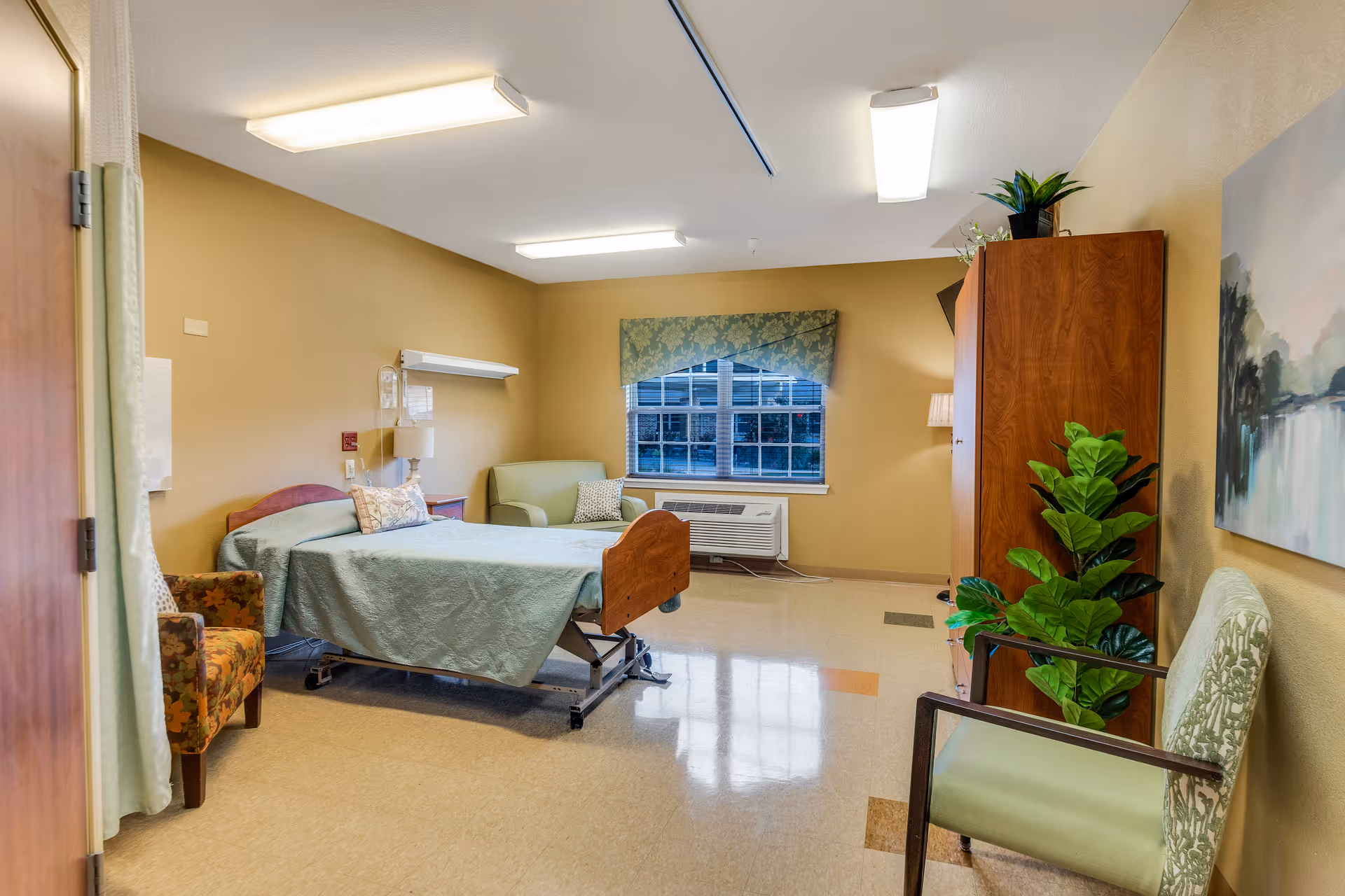 A well-lit senior living bedroom with a hospital-style bed covered in a green bedspread, a floral armchair, a green armchair, a wooden cabinet, a potted plant, a window with a green valance, and a landscape painting on the wall.
