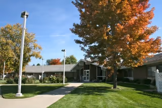 Exterior view of a single-story building with a gray roof and beige walls, surrounded by green grass and trees with autumn-colored leaves under a clear blue sky.