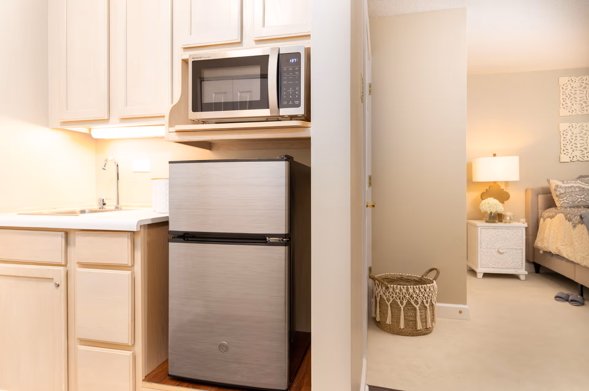 A small kitchen area with light wood cabinets, a stainless steel mini refrigerator, and a microwave oven mounted above it. To the left is a countertop with a sink and a faucet. To the right, an open doorway leads to a bedroom with a nightstand, a lamp, decorative wall art, and a bed with pillows and a blanket. A woven basket is placed on the floor near the doorway.