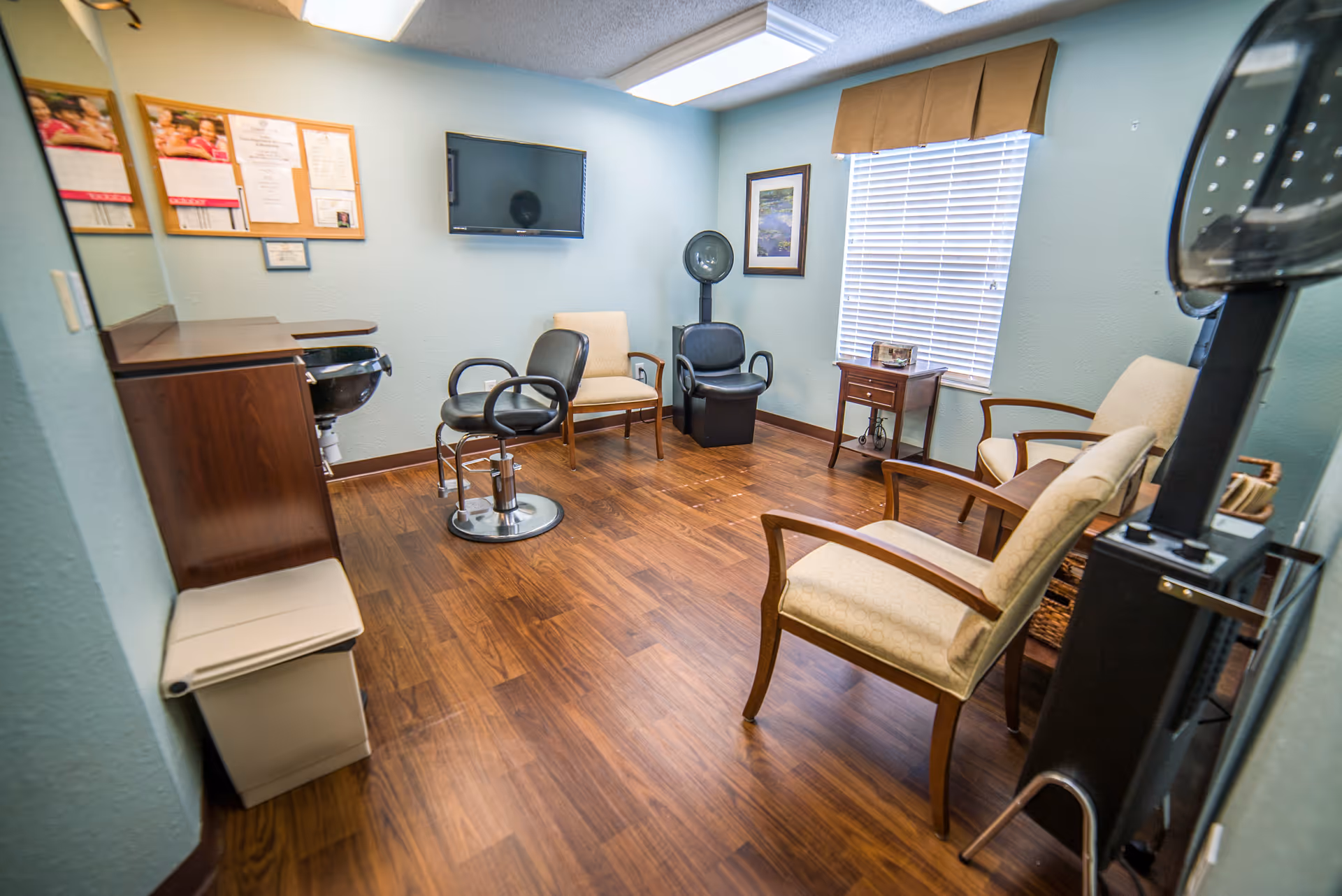 A small salon room with light blue walls and wood flooring. The room contains a black salon chair, a hair dryer chair, three beige armchairs, a small wooden side table, a wall-mounted TV, a bulletin board with papers, and a window with blinds and a brown valance.