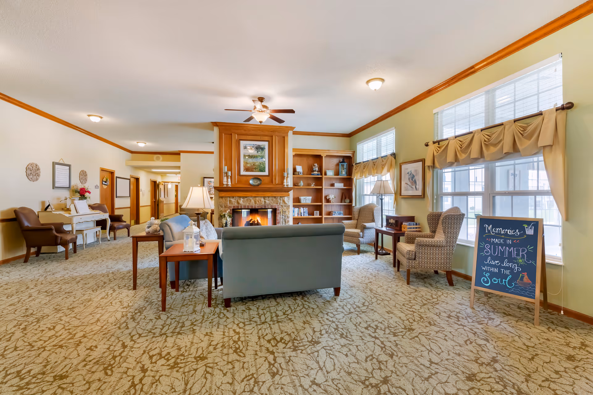 A cozy living room area in a senior living facility with a fireplace in the center, flanked by built-in wooden shelves. The room has comfortable seating including a sofa and armchairs, a ceiling fan, large windows with valances, and a decorative sign that reads 'Memories made in summer live long within the soul.' The walls are painted light green and beige with wooden trim, and there is a carpet with a leaf pattern.