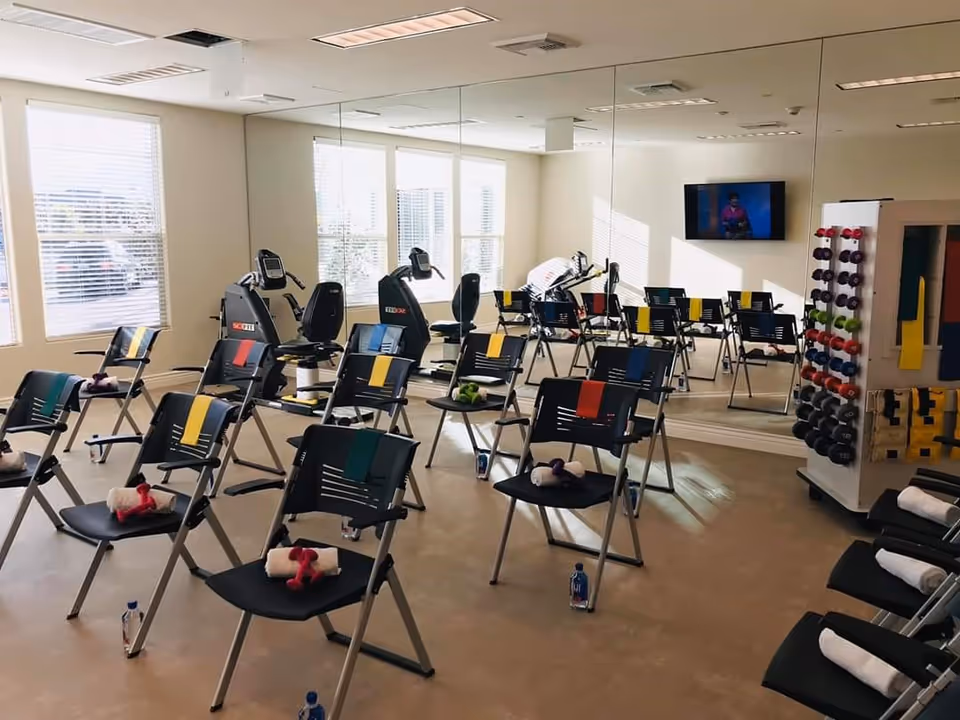 A fitness room with multiple black chairs arranged in rows, each chair holding a rolled towel and a pair of small dumbbells. Resistance bands are draped over the backs of the chairs. The room has large windows letting in natural light, a wall covered with mirrors, exercise bikes, a TV mounted on the wall, and a rack filled with various colored dumbbells and exercise bands.