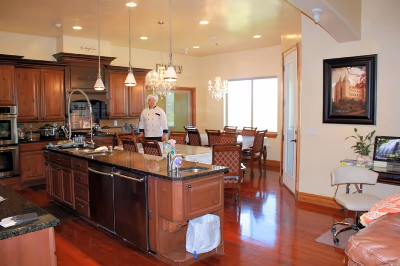 A spacious kitchen and dining area with wooden cabinets, a large island with a sink, and granite countertops. A chef in a white uniform stands near the dining table with multiple chairs. The room has hardwood floors, pendant lights, chandeliers, and a window letting in natural light. There is also a small desk area with a computer and a chair on the right side.