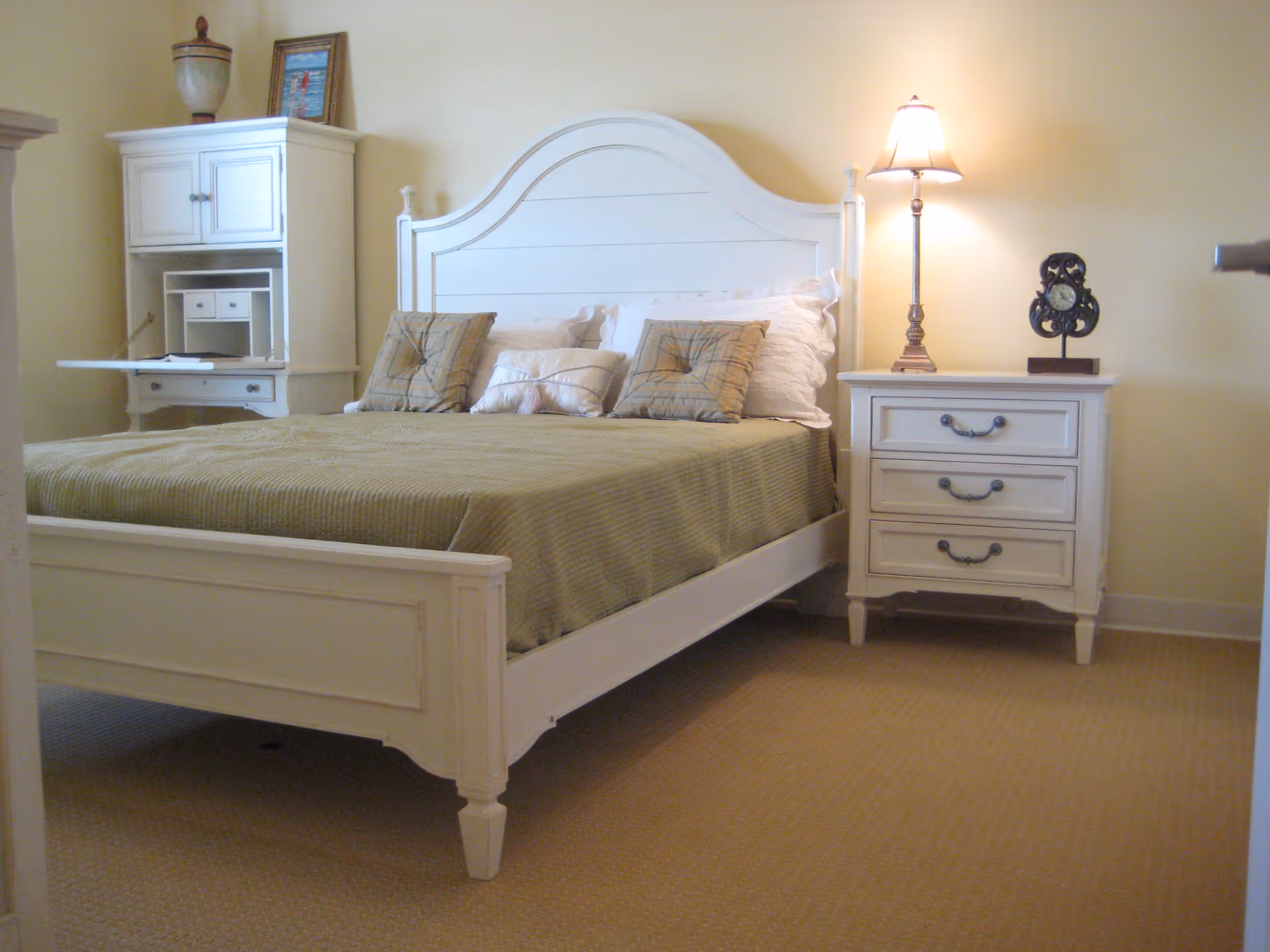 A bright bedroom with a white wooden bed dressed in a green coverlet, a nightstand with a lamp and a small cabinet against a pale yellow wall.