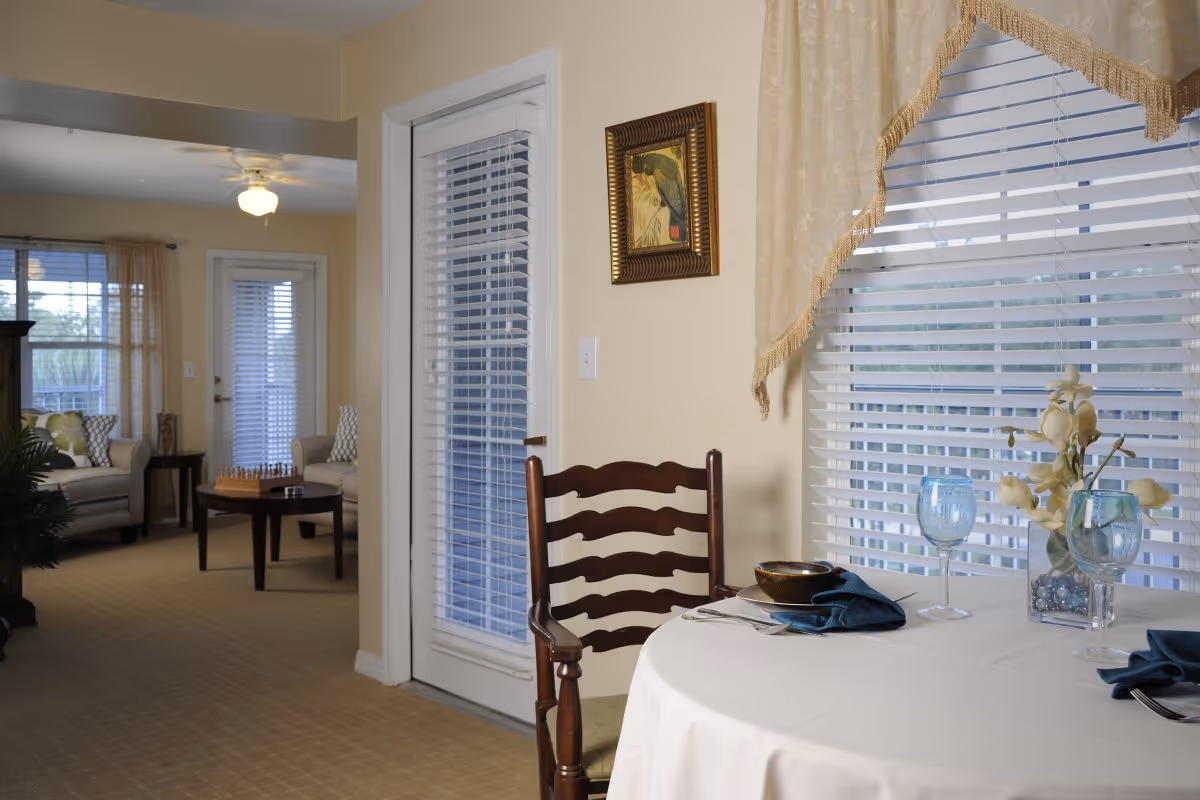 A dining table set with glassware and napkins by a window with blinds, adjacent to a living room visible through an open doorway.